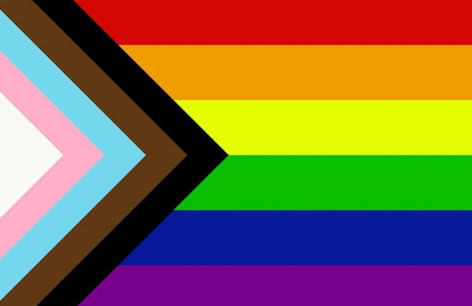 Progress pride flag, with horizontal red, orange, yellow, green, blue and purple stripes and a chevron shape on the left side with white, pink and blue stripes
