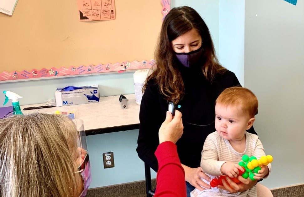 A CHEO audiology assessment taking place, with a baby in their parent's lap and a staff member holding up a microphone