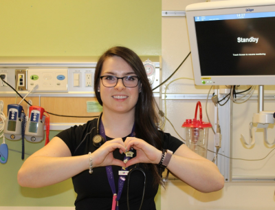 Molly, a CHEO nurse, wearing normal scrubs and smiling and making a heart with her hands