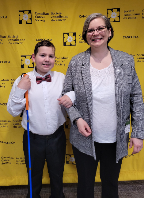 Ollie and Dawn pose in front of the Canadian Cancer Society yellow backdrop on their Day on the Hill. Ollie wears a white shirt, red bow tie and hold his mobility cane. Dawn wears glasses and her grey blazer.